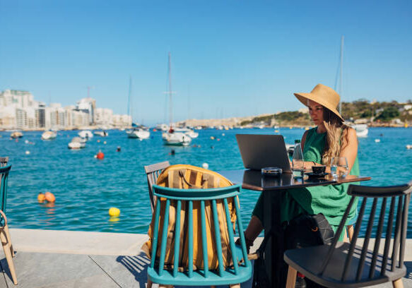 A woman with a straw hat on her head works remotely sitting in a coffee shop not far from the sea